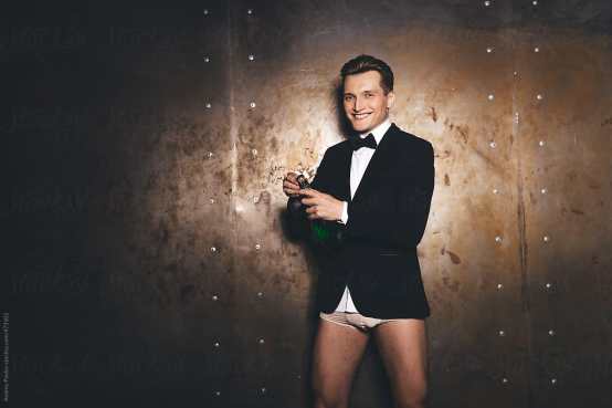 A young white man wearing a tuxedo jacket, white shirt, black bow tie, white underwear, and a smile. He's opening a bottle of champagne because of course he is.