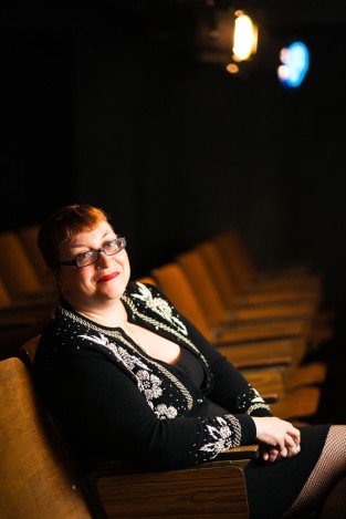 A smiling redhead wearing a black beaded sweater, a black dress, black-framed glasses, and red lipstick sits in a theater with her hands folded in her lap.